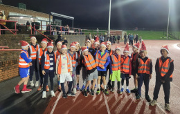 Group of Highgate Harriers U11 junior runners wearing Santa hats and smiling during a festive evening training session at the athletics track.